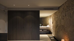 Een moderne slaapkamer met een groot bed, een houten garderobe, een accentmuur van steen en een badkamer met een wastafel en kaarsen.