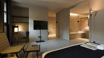 Una habitación de hotel moderna con una cama, un escritorio con televisor de pantalla plana, una silla, una mesita con un florero y vasos, y un baño con lavamanos y espejo.