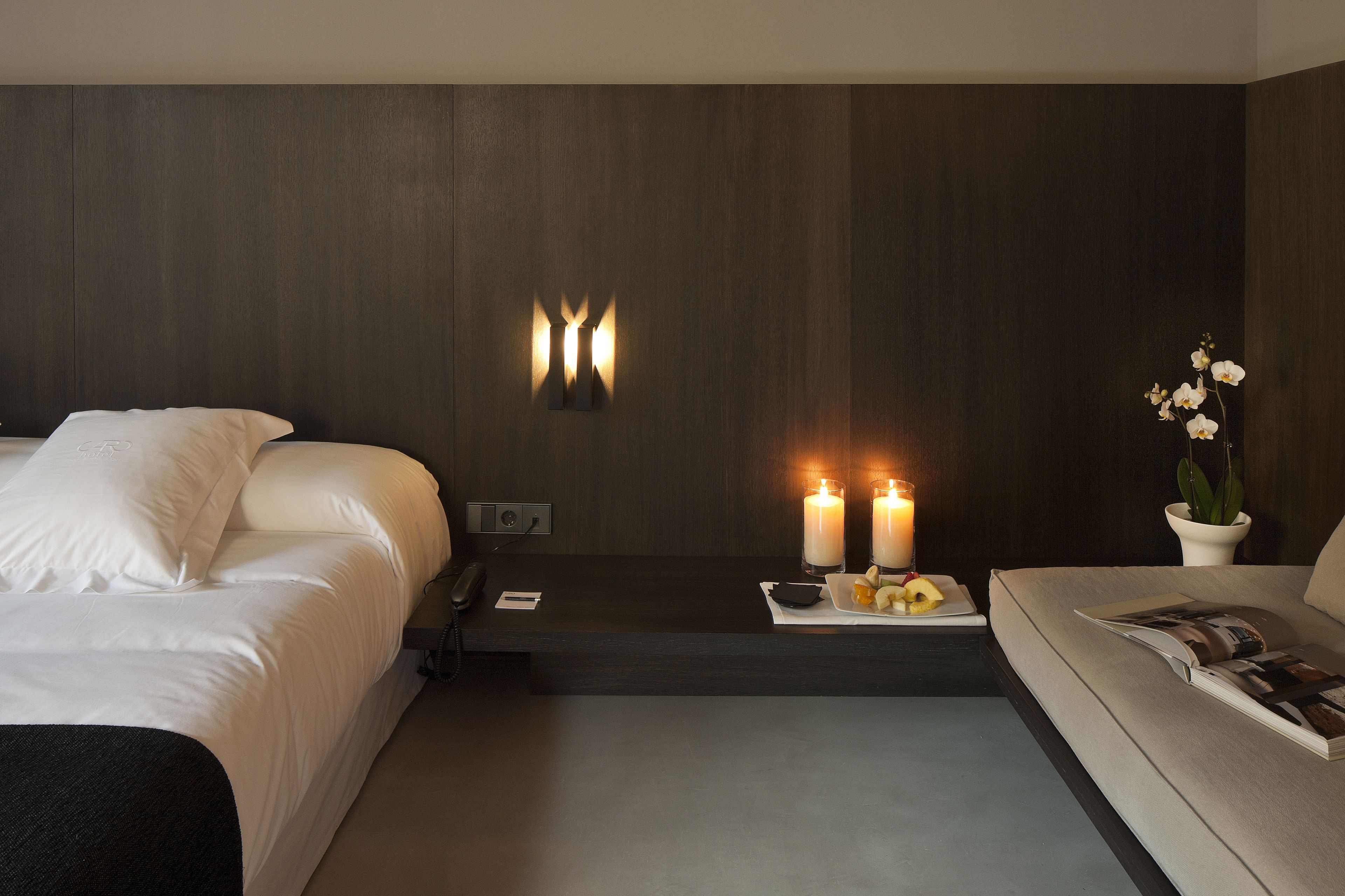 Una habitación de hotel moderna con una cama, un sofá, una mesita de noche con teléfono, y una lámpara de pared.