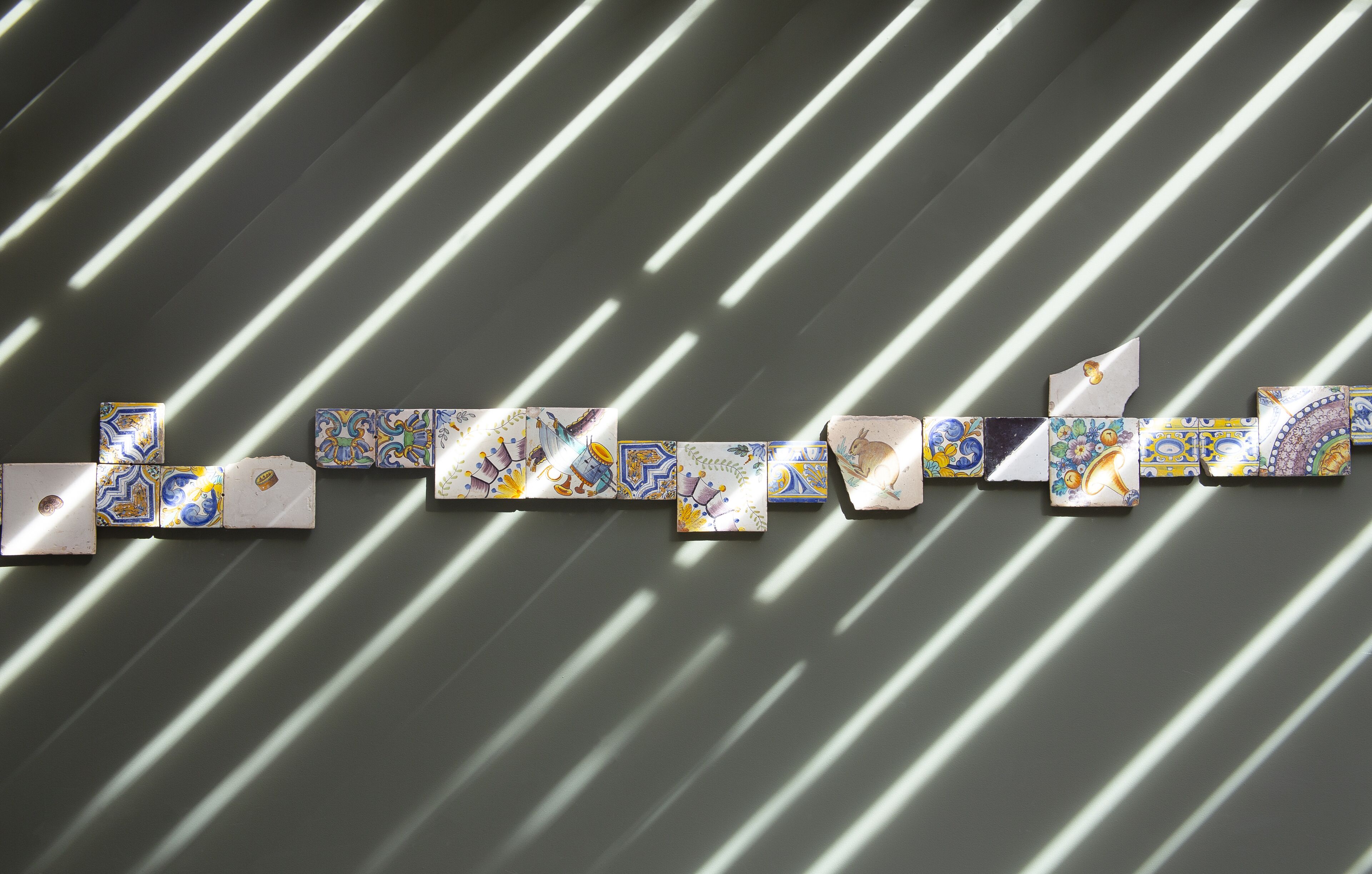 Una serie de baldosas con patrones geométricos dispuestas en una secuencia lineal frente a una pared con sombras diagonales.