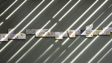 Una serie de baldosas con patrones geométricos dispuestas en una secuencia lineal frente a una pared con sombras diagonales.