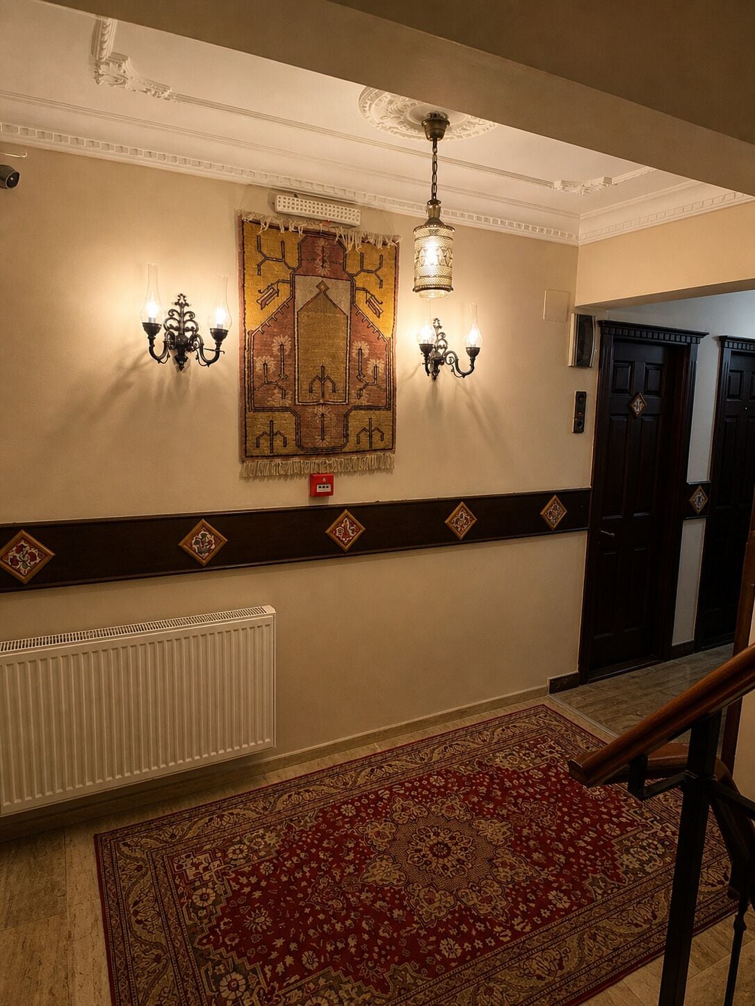Un couloir avec un mur orné de motifs, un radiateur, une moquette rouge et un escalier.