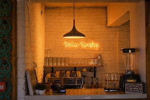 Una zona de bar con un letrero de neón que dice 'Bella Rooftop', una máquina de café, una licuadora y una caja para propinas sobre la barra.