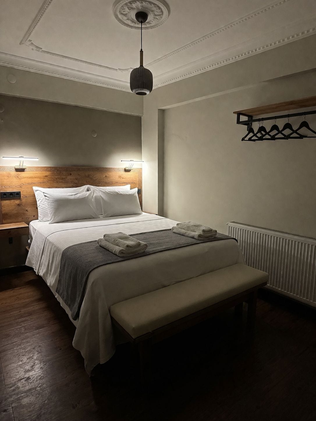 Une chambre d’hôtel comprenant un lit, des tables de chevet, un banc, un luminaire suspendu et un radiateur.