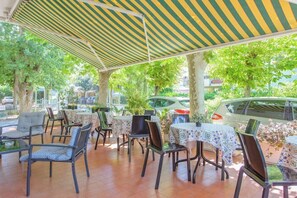 Un café al aire libre con mesas y sillas, cubierto por toldos a rayas, rodeado de árboles y coches aparcados.