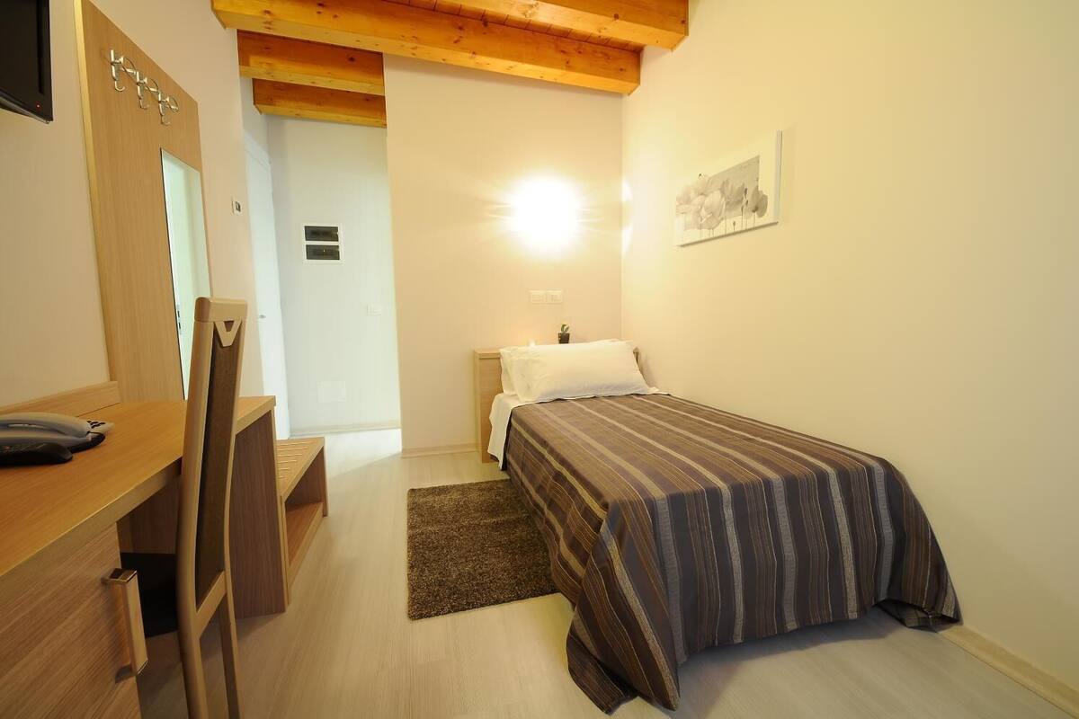 A bedroom with a bed, bedside table, chair, desk, and a small bedside lamp.