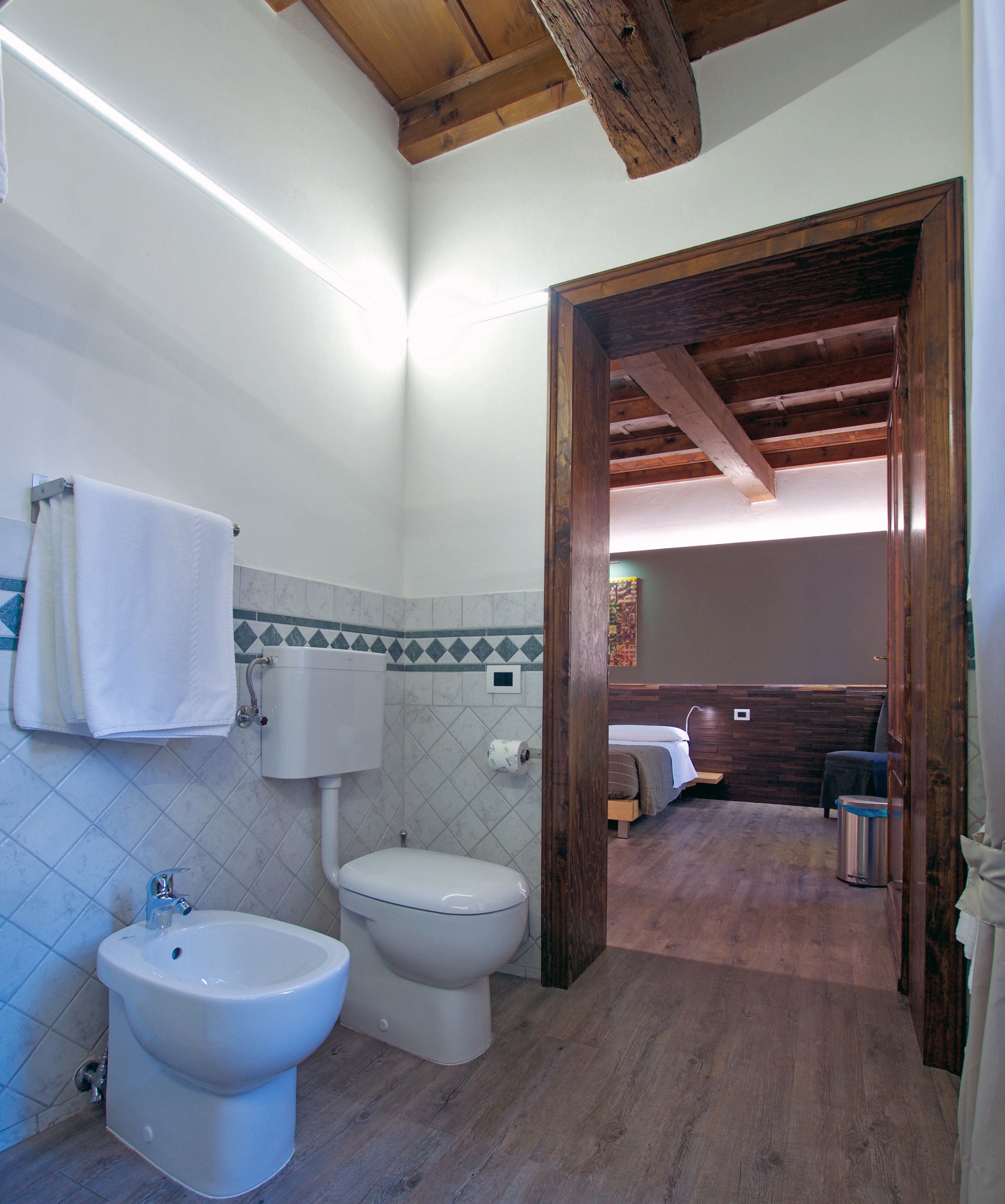 A bathroom with a bidet, toilet, and a sink. There is a wooden ceiling and a doorway leading to a bedroom.
