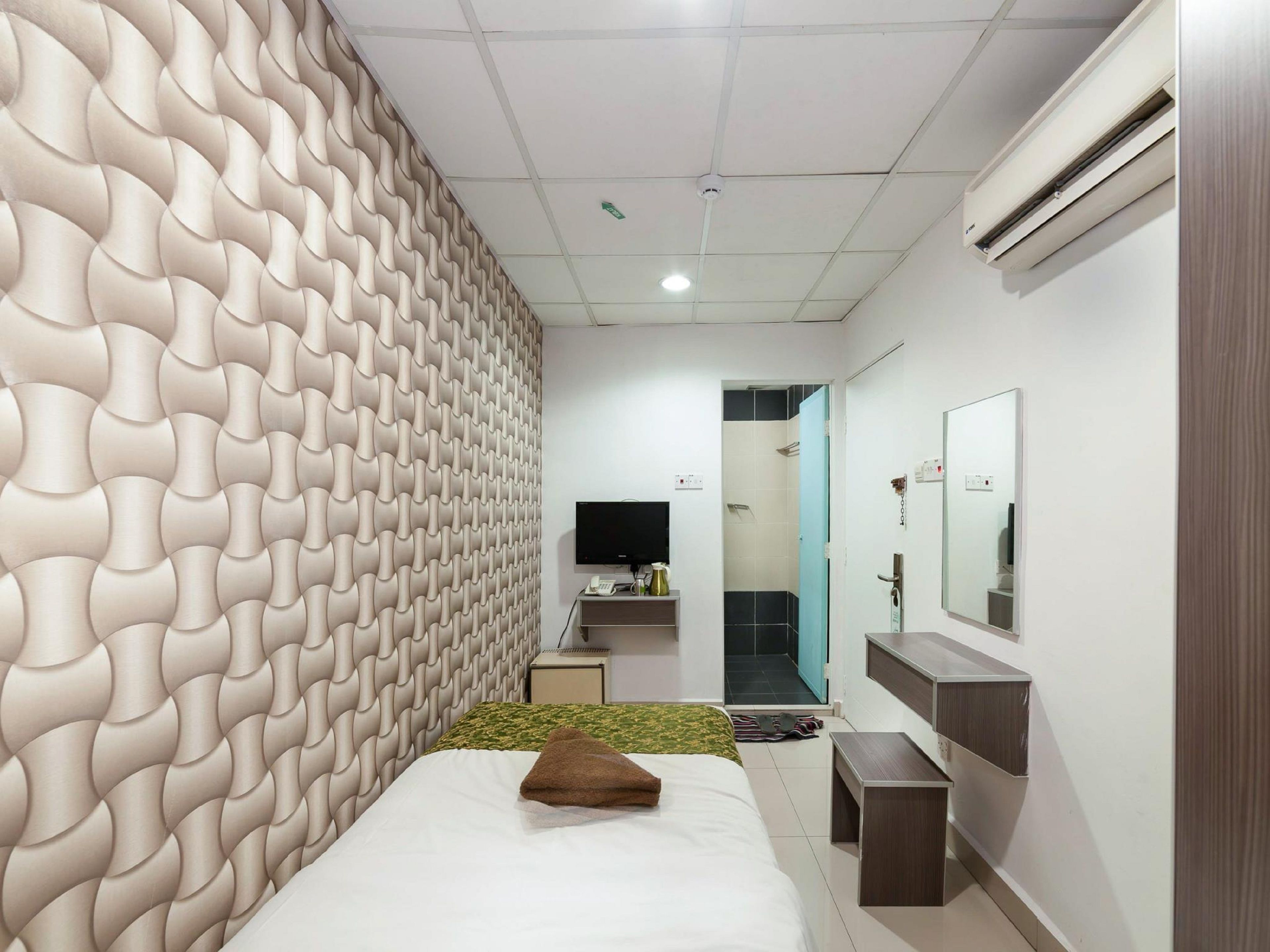 Una habitación de hotel moderna con una pared de textura única, una cama con colcha verde y blanca estampada, una mesita de noche pequeña, un espejo y un televisor montado en la pared.