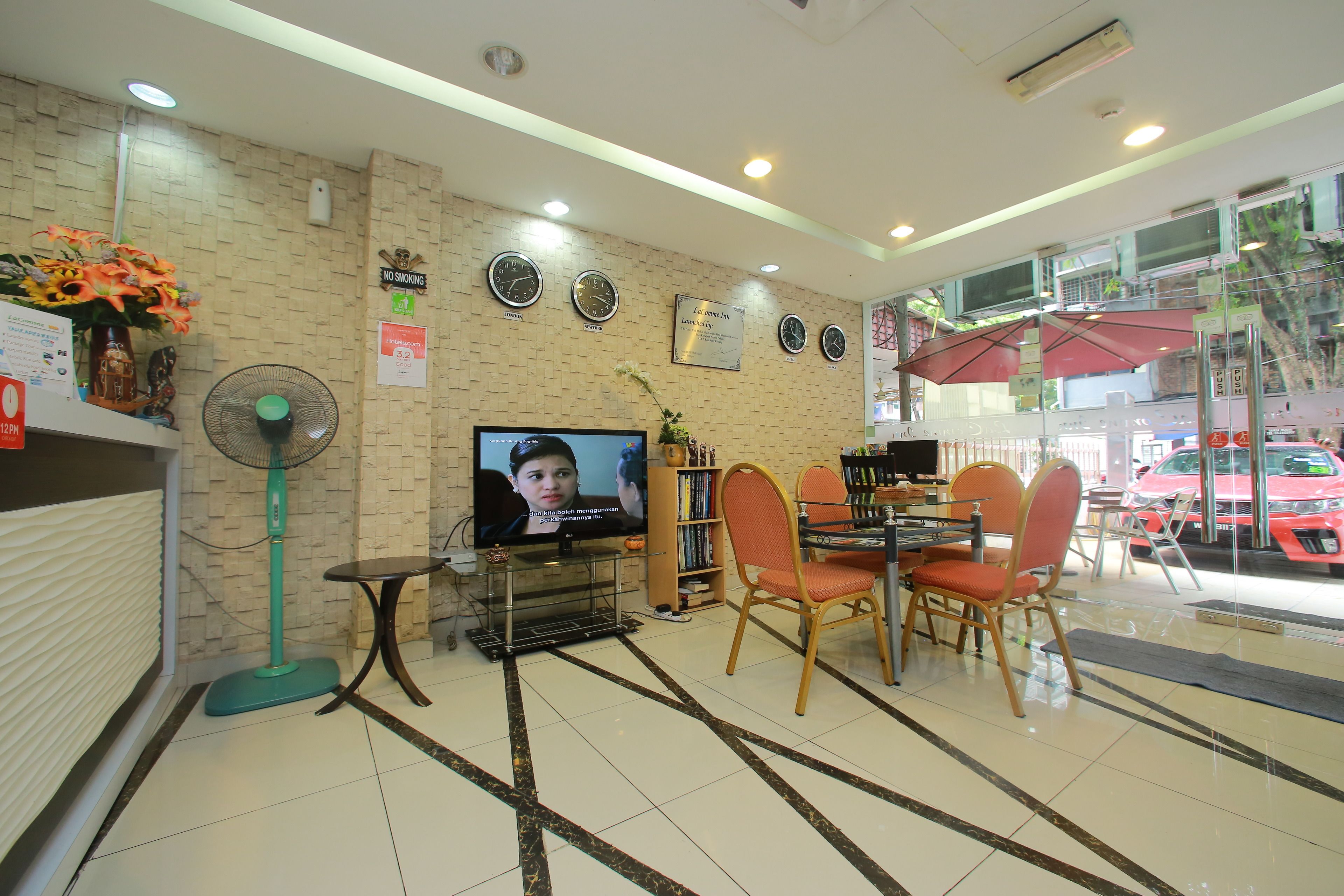 Un hall de hotel con mostrador de recepción, pantalla de televisión, ventilador y zona de comedor con sillas y mesas.