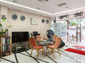 Un vestíbulo con pared de ladrillo, relojes, televisor, mesa de vidrio con sillas y alfombra roja.
