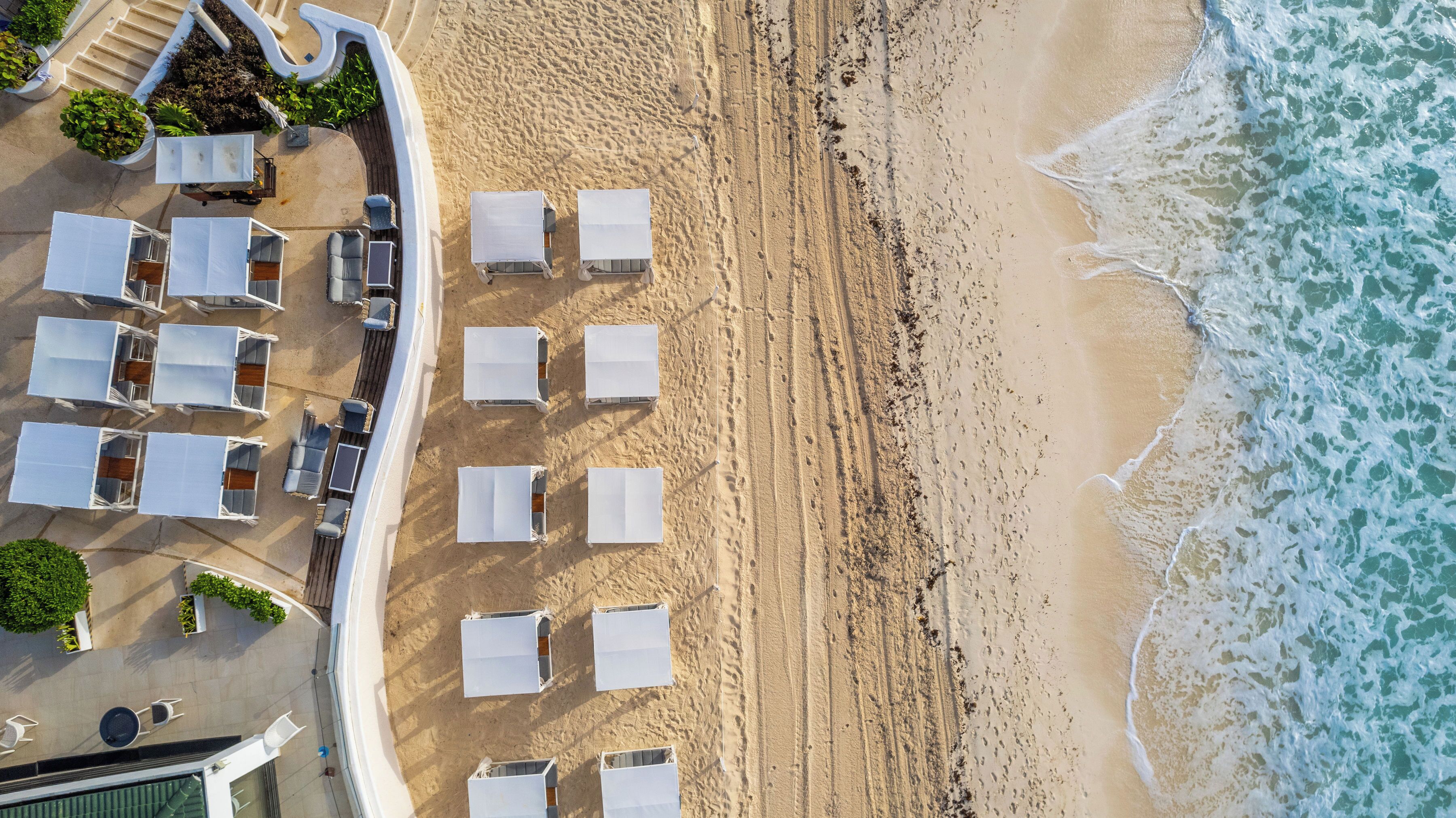 An aerial view of a beachside hotel with rows of sun loungers and umbrellas, a sandy beach, and ocean waves.