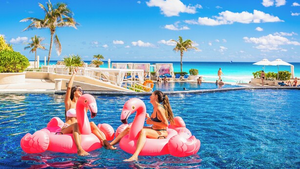 A pool area with a clear view of the ocean, lounge chairs, and palm trees.