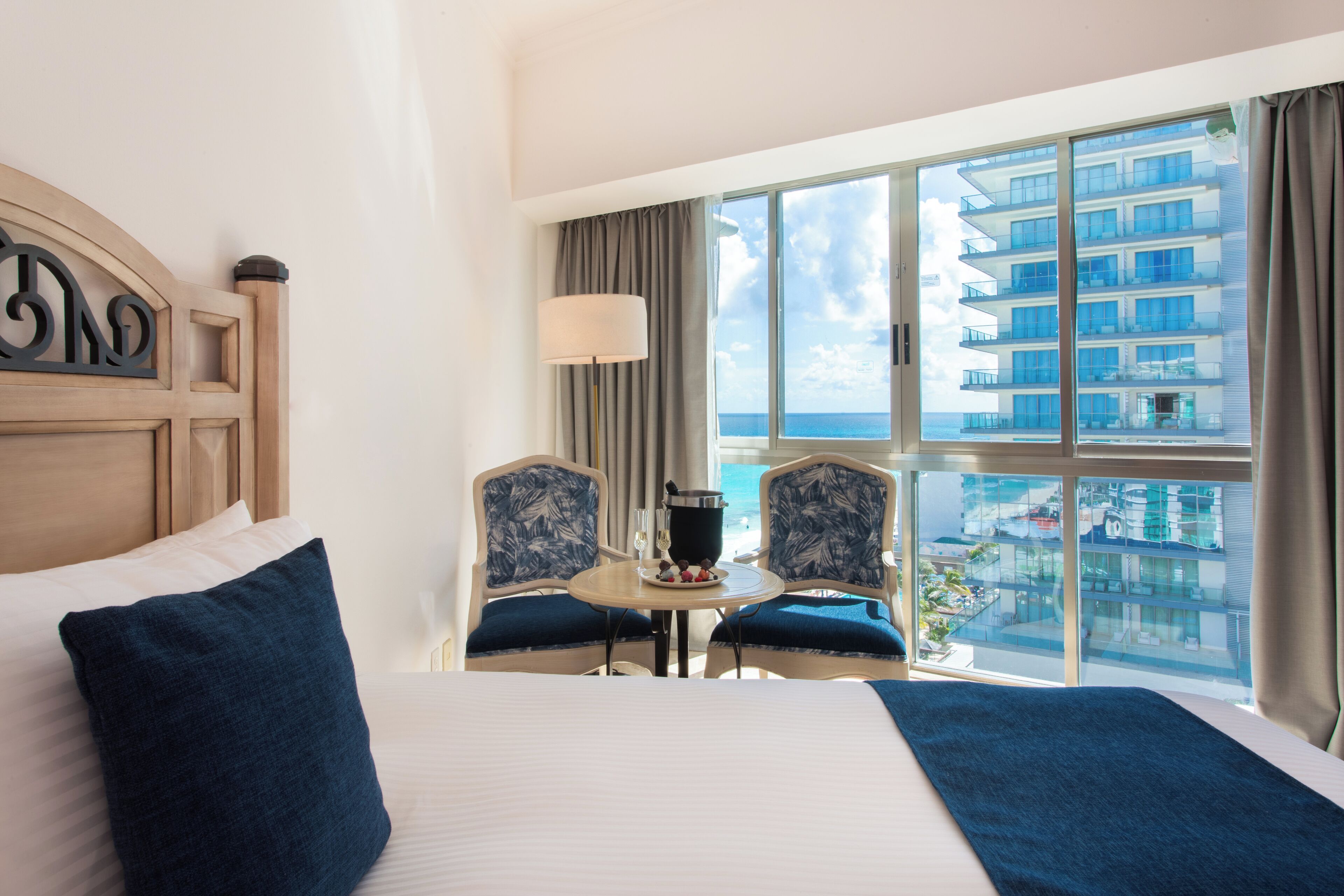 Une chambre d’hôtel avec un grand lit, deux fauteuils, une petite table avec une boisson, et une vue sur l’océan et les bâtiments.