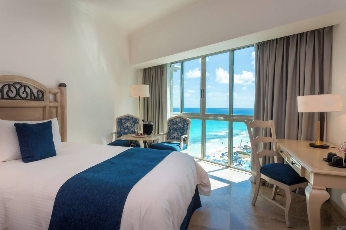 A hotel room with a large bed, two chairs, a desk, and a view of the beach.