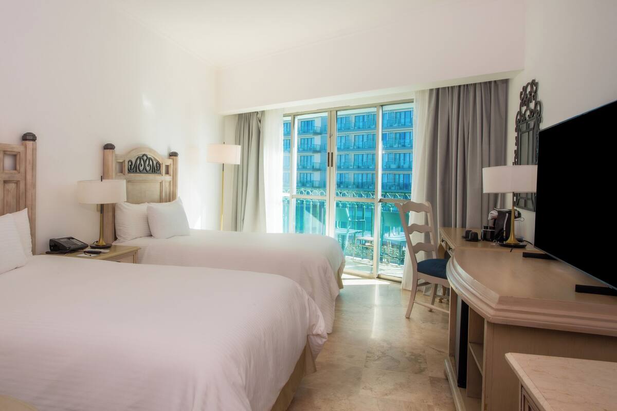 A hotel room with two beds, a desk, a chair, a TV, and a balcony with a view of buildings.