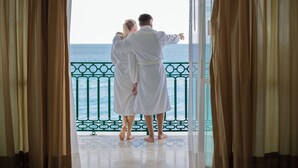 A couple in white robes standing at a balcony with a view of the sea.