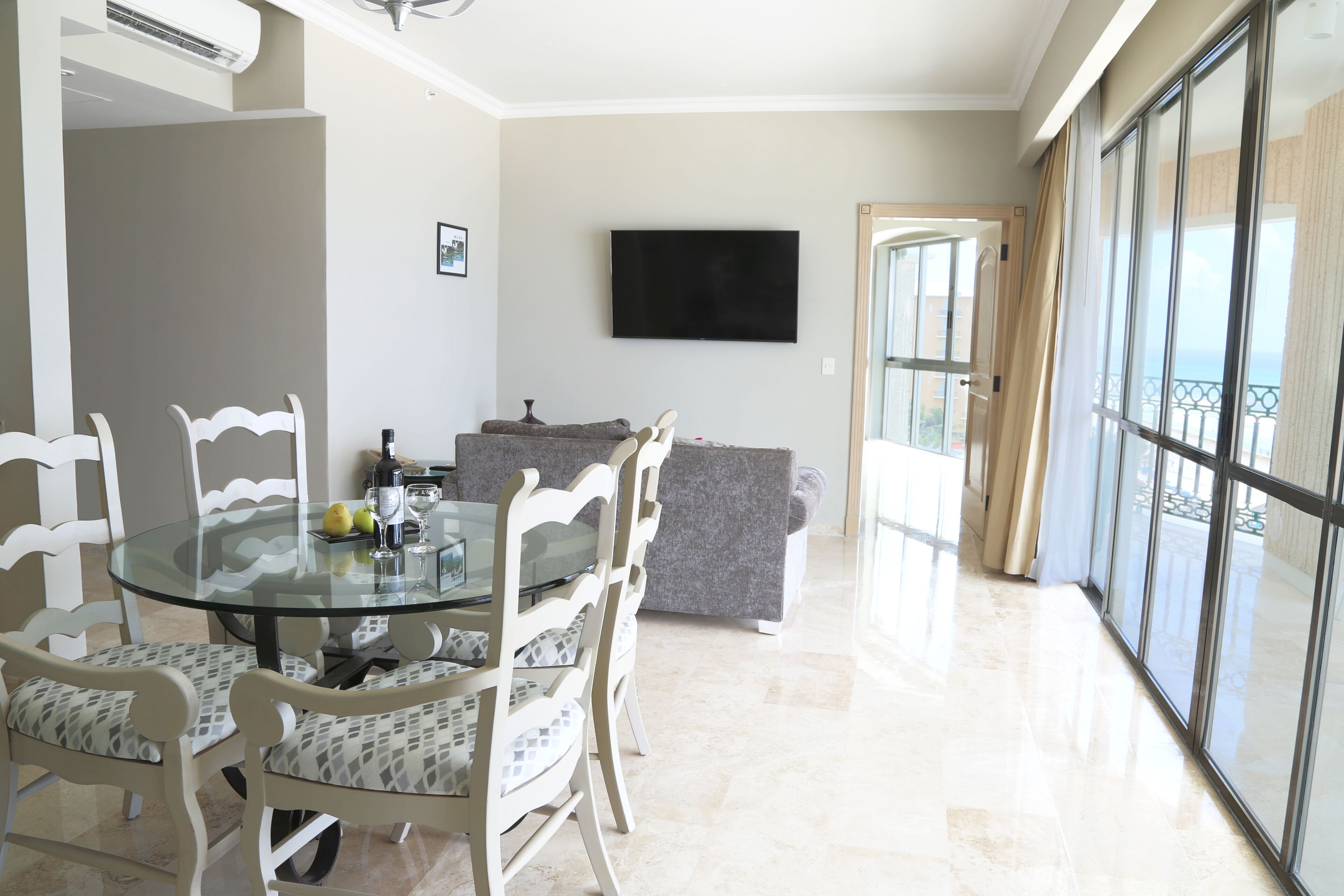 Un comedor con una mesa y sillas de vidrio, un televisor de pantalla plana en la pared, y vistas al balcón exterior a través de amplias ventanas.
