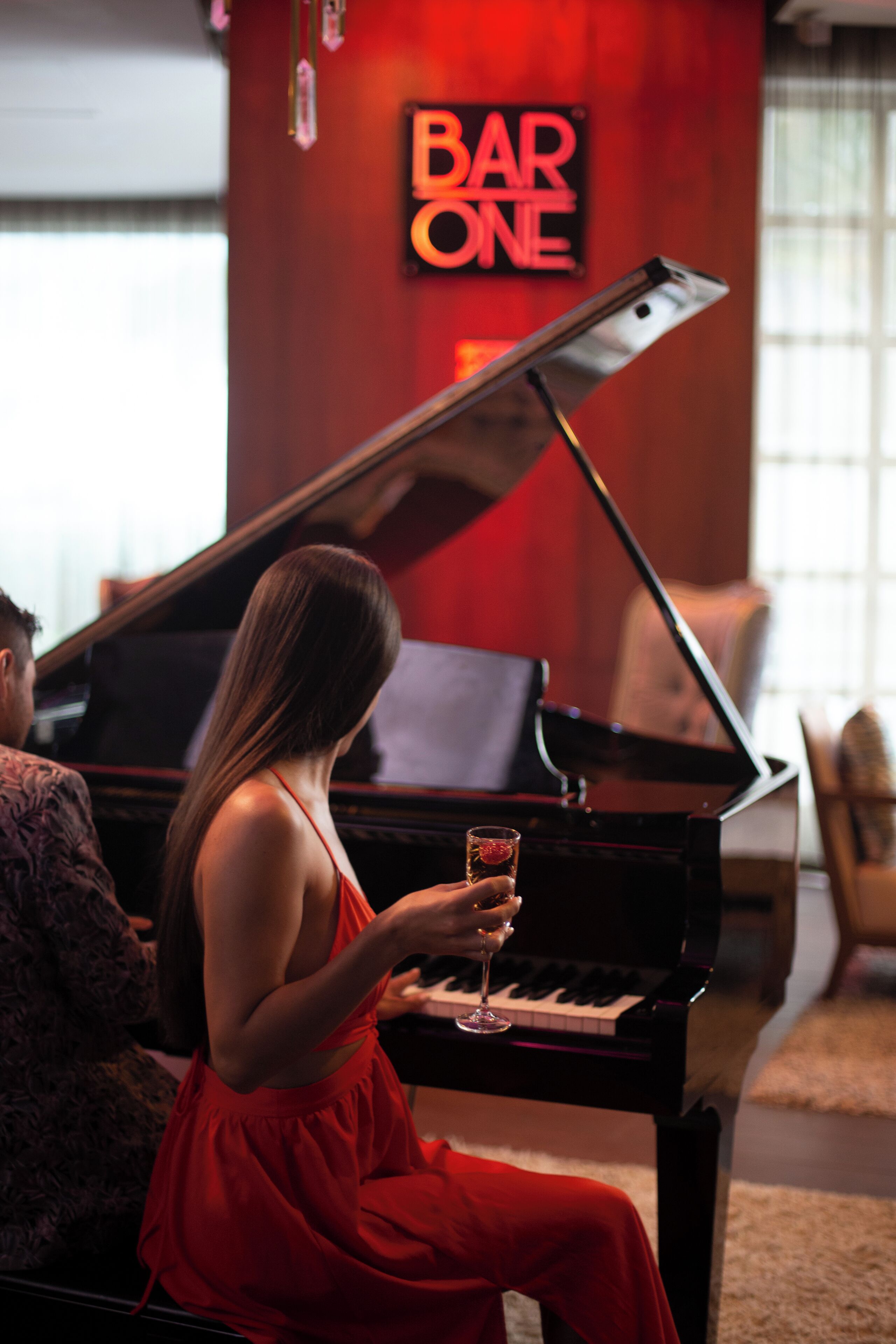 A woman in a red dress sitting at a grand piano in a room with a 'BAR ONE' sign on the wall.