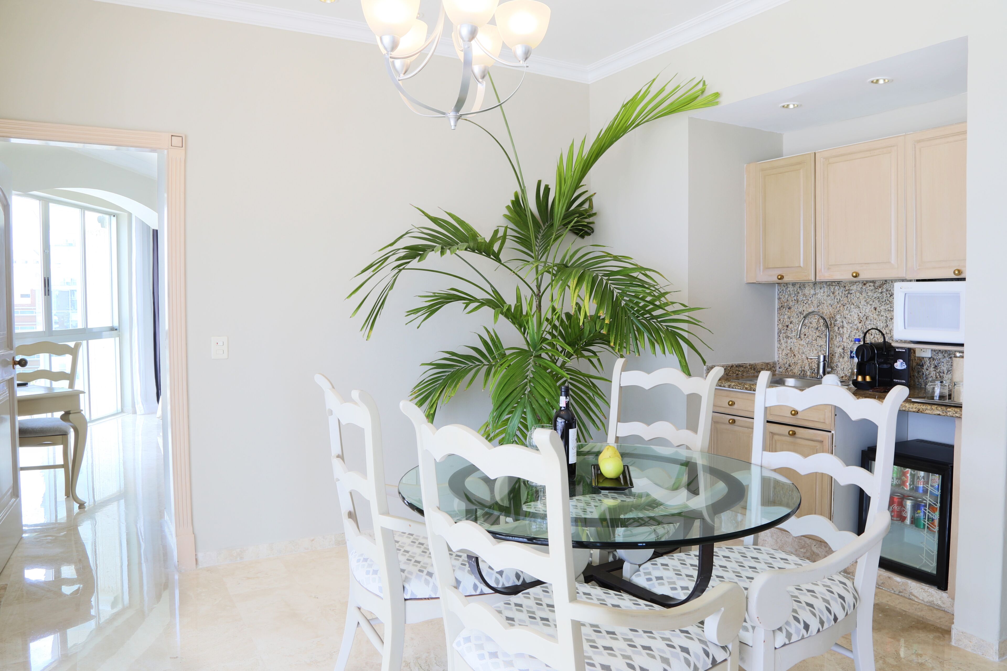 A dining area with a glass table surrounded by white chairs, a potted plant, and a kitchen in the background.