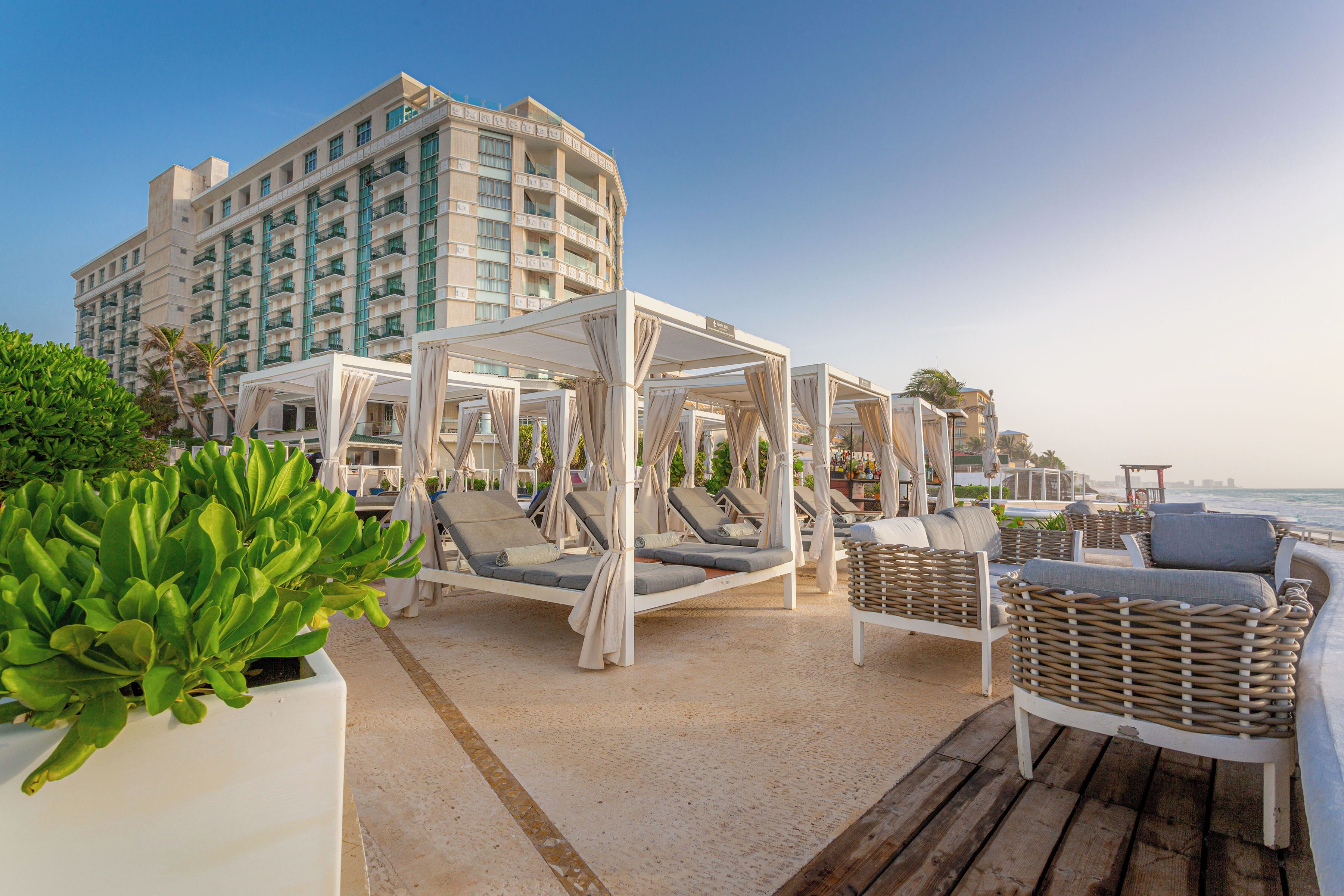 A beachside hotel with outdoor seating areas, including white canopies and wicker furniture, overlooking the ocean.