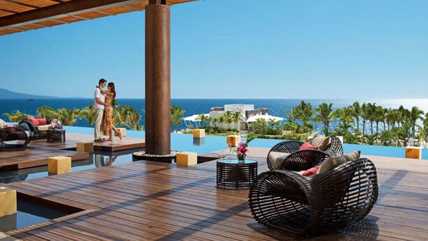 A couple on a wooden deck overlooking a pool with lounge chairs and a view of the ocean and resort buildings.