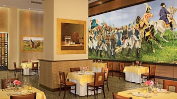 Un comedor con mesas dispuestas para comidas, un cuadro histórico grande en la pared y una estantería con libros.