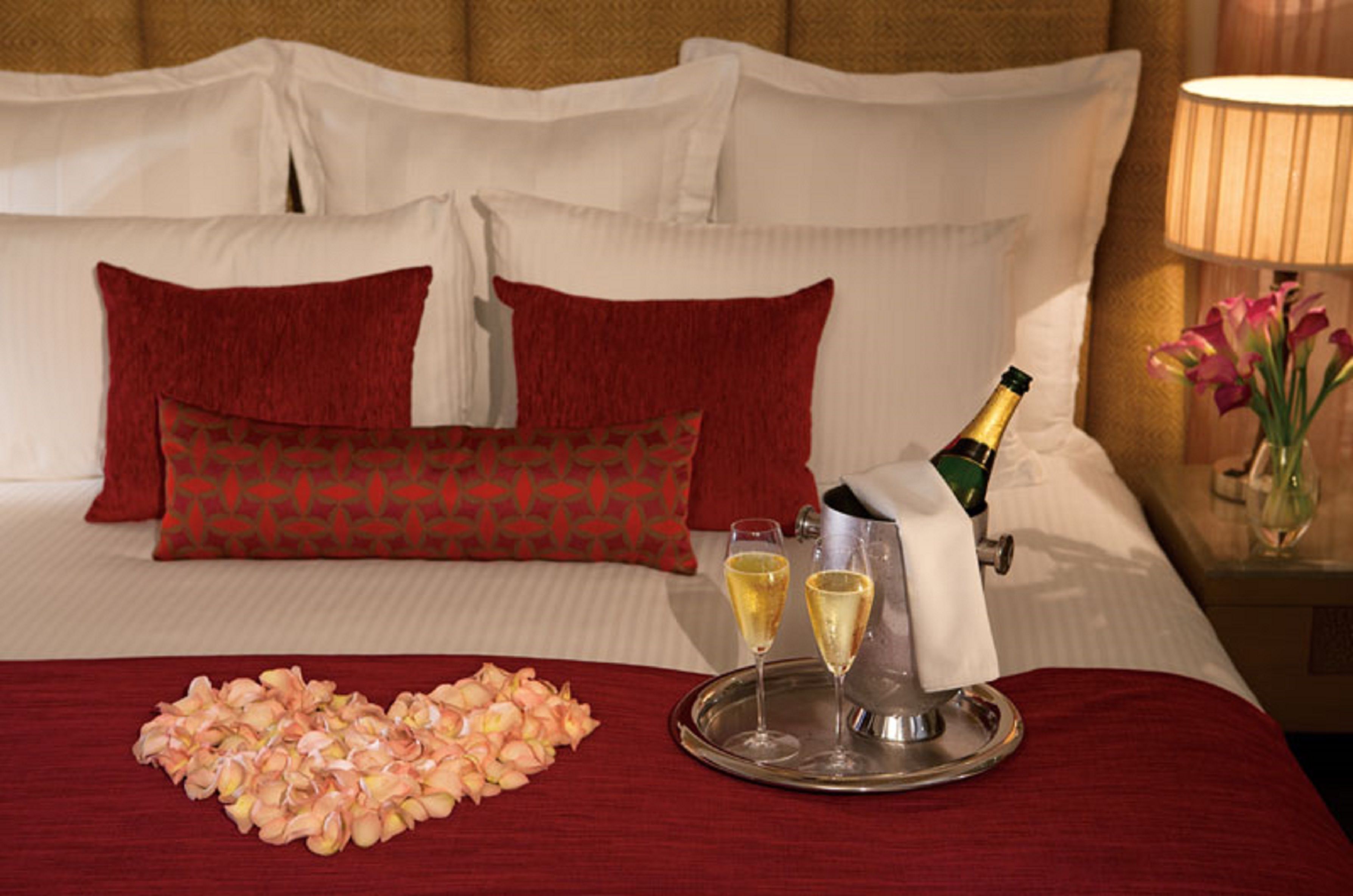 A neatly made bed with red and white bedding, a tray with champagne glasses, and a bouquet of flowers.