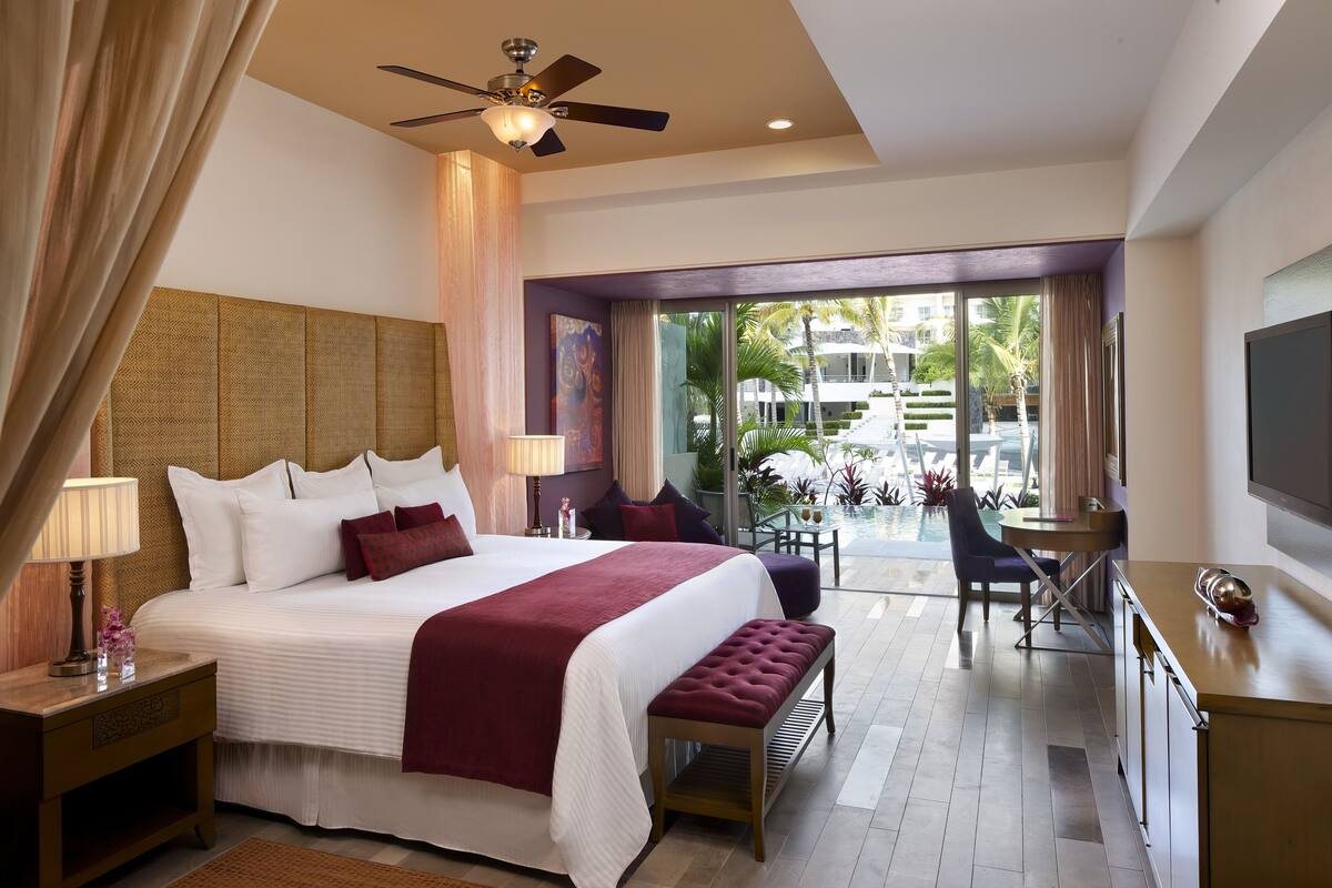 Ein modernes Hotelzimmer mit einem großen Bett, einem Deckenventilator, einem Schreibtisch und Blick auf einen Pool und Palmen.