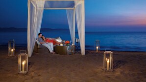 A beach setting with a canopy and a couple relaxing on a bed. There are lanterns and a table with a drink.
