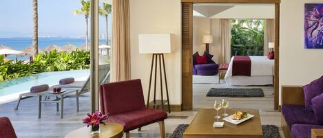 Habitación de hotel con vista al mar, una cama, una mesa de comedor con un plato de comida y un área de descanso con una silla y una mesita.