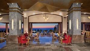 A spacious lobby with a view of the ocean at sunset, featuring a central seating area with red chairs and a chandelier.