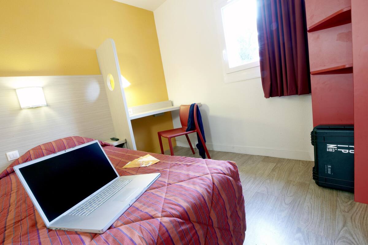 Quarto de hotel com uma cama, escrivaninha, cadeira, mala e janela com cortinas.