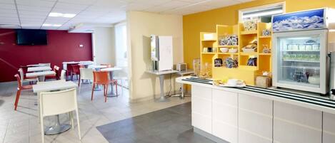 Una cafetería moderna con mesas y sillas blancas, un mostrador amarillo con dispensadores de comida y bebida, y un refrigerador blanco.