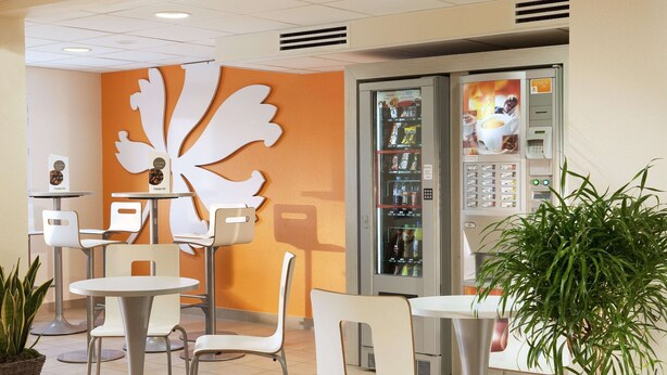 A modern cafe with white chairs and tables, a large decorative wall piece, and a beverage vending machine. - Hotel Premiere Classe Pau EST Bizanos (Bizanos)