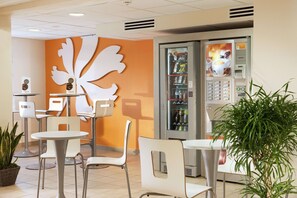 A modern cafe with white chairs and tables, a large decorative wall piece, and a beverage vending machine. - Hotel Premiere Classe Pau EST Bizanos (Bizanos)