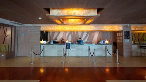 A hotel reception area with a modern design, featuring a reception desk, a digital information board, and a wooden floor.