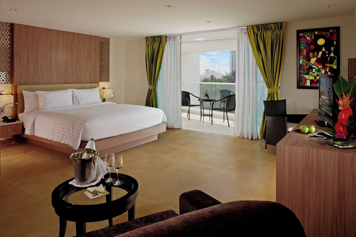 Une chambre d’hôtel avec un grand lit, un bureau avec un ordinateur, un balcon avec des chaises et une vue sur la ville.