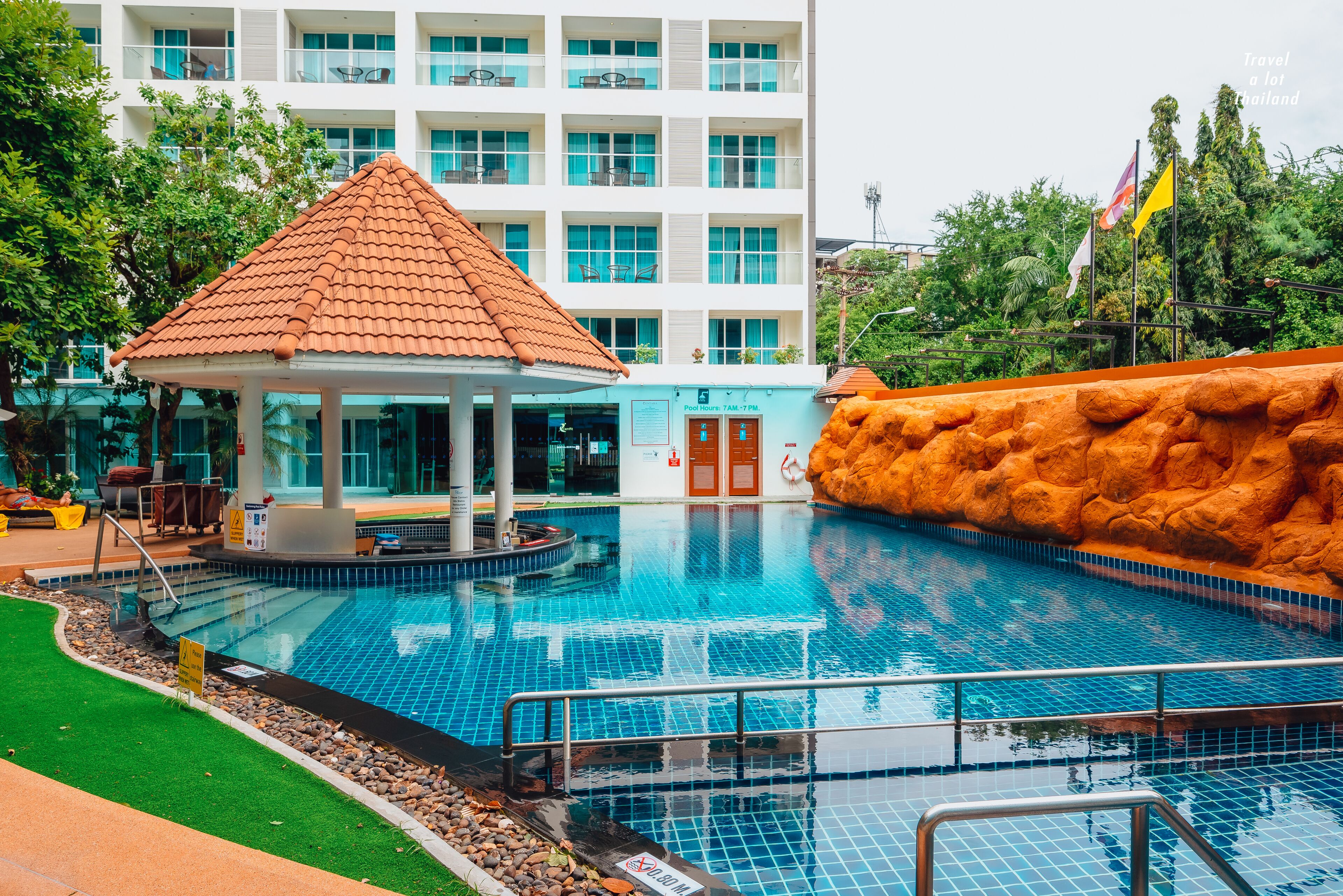 A swimming pool with a tiled deck, a covered seating area, and a building with multiple windows in the background.