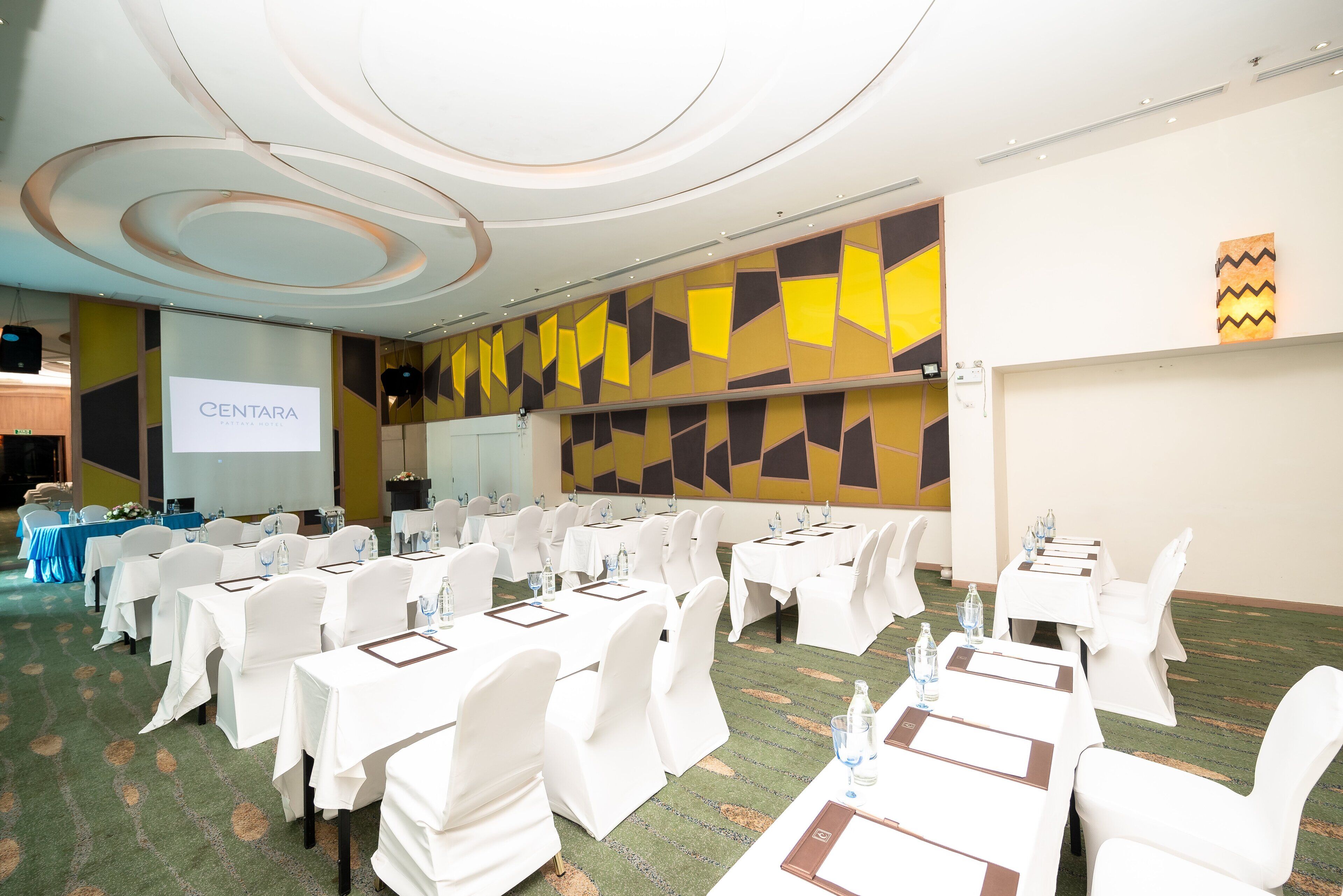 A conference room with white tables and chairs, a projector screen, and a colorful geometric wall design.