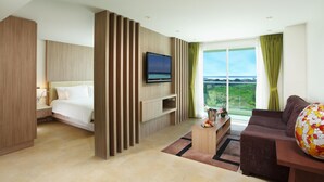 A modern hotel room with a bed, a TV mounted on the wall, a sofa, and a large window offering a view of the outdoors.