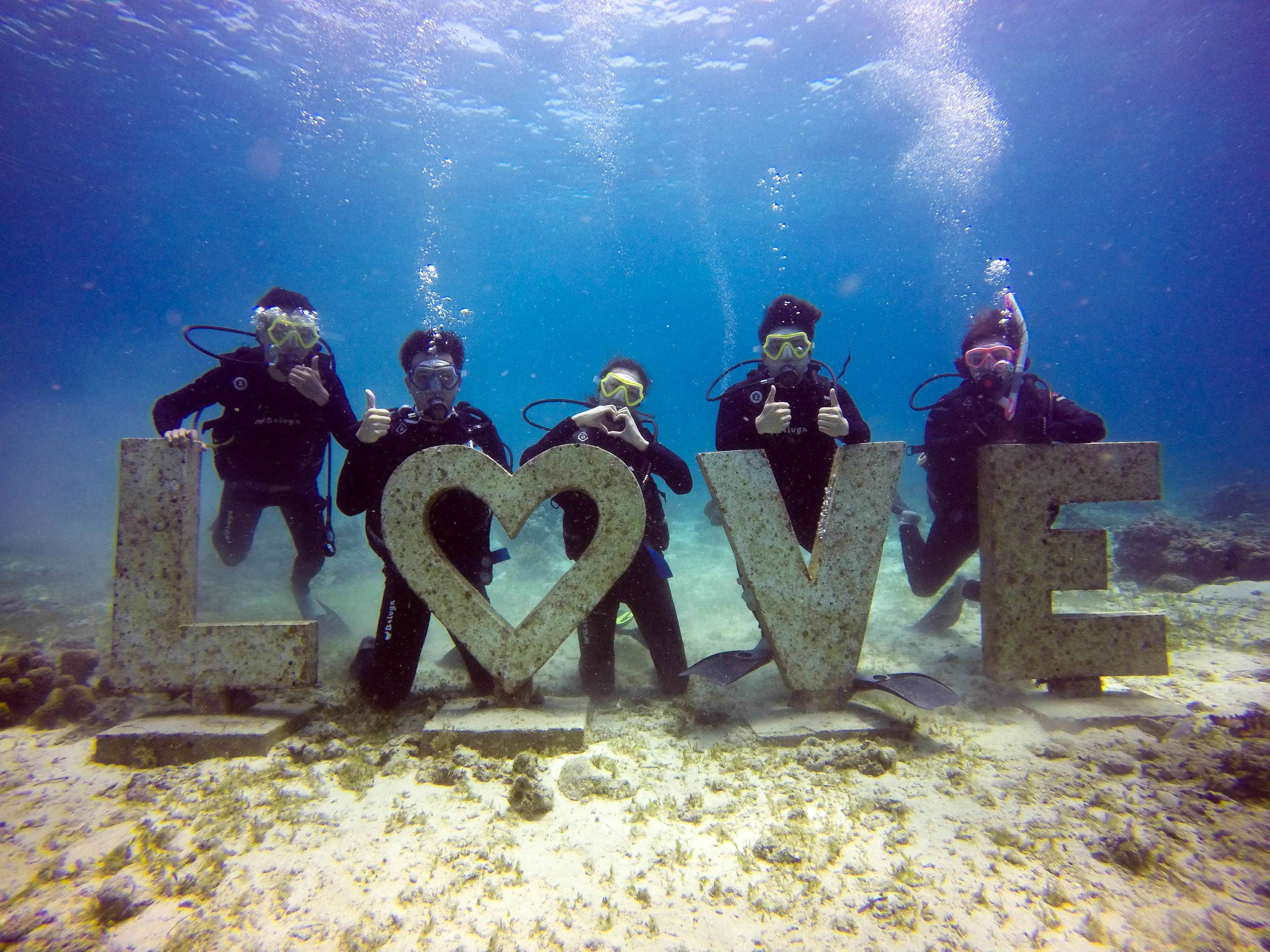 Four divers holding large letters spelling "LOVE" underwater.