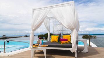 A white canopy bed with colorful pillows by a poolside, a table with snacks, and a view of the sea.