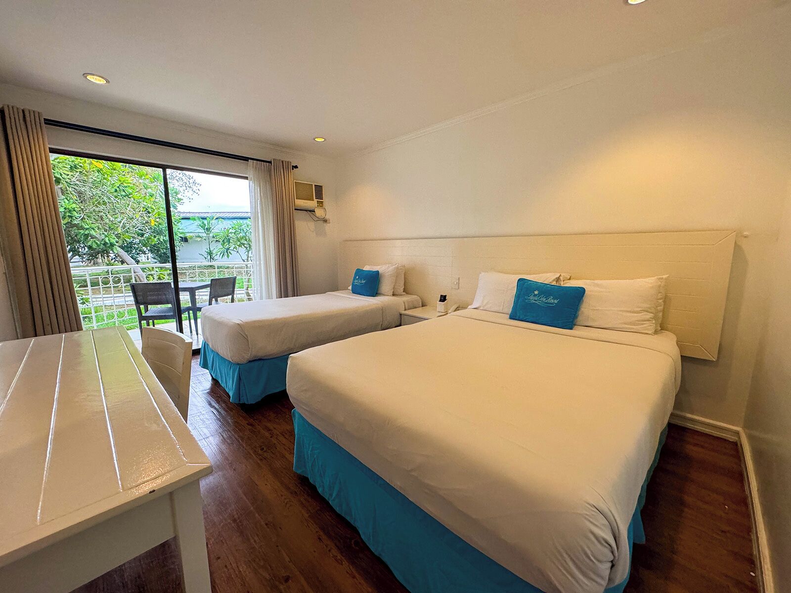 A hotel room with two beds, a balcony with a table and chairs, and a view of greenery outside.
