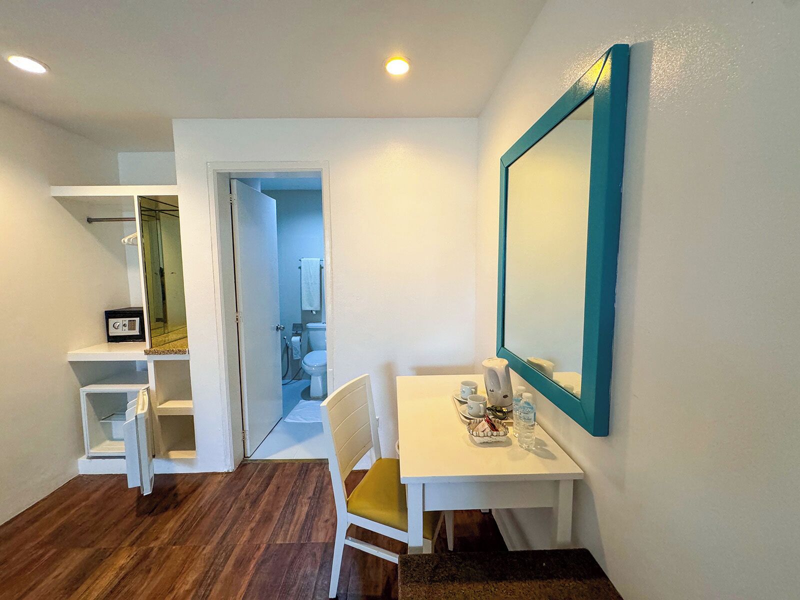 Una habitación de hotel con un escritorio blanco, una silla con cojín amarillo, un espejo grande con marco azul turquesa y suelo de madera.