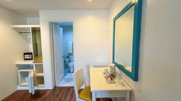 Una habitación de hotel con un escritorio blanco, una silla con cojín amarillo, un espejo grande con marco azul turquesa y suelo de madera.
