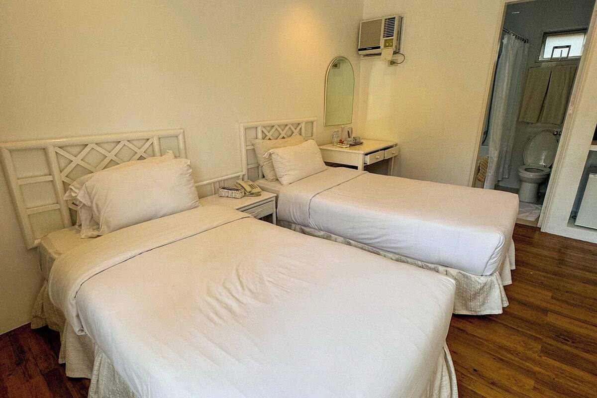 A room with two beds, a bedside table with a lamp, a mirror, and an air conditioning unit.