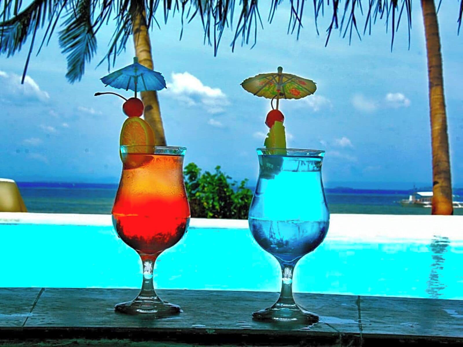 Two drinks with umbrellas on a wooden surface by a pool with a view of the ocean.