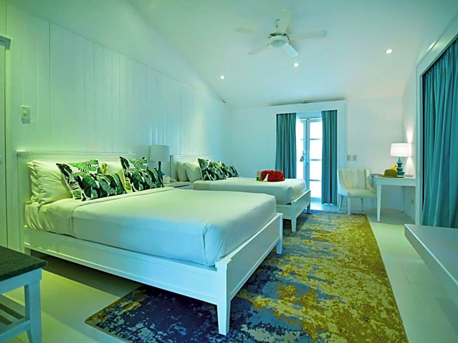 Une chambre d’hôtel avec un grand lit, deux petits lits, un bureau, une chaise, un ventilateur de plafond et une vue sur l’extérieur par une porte.