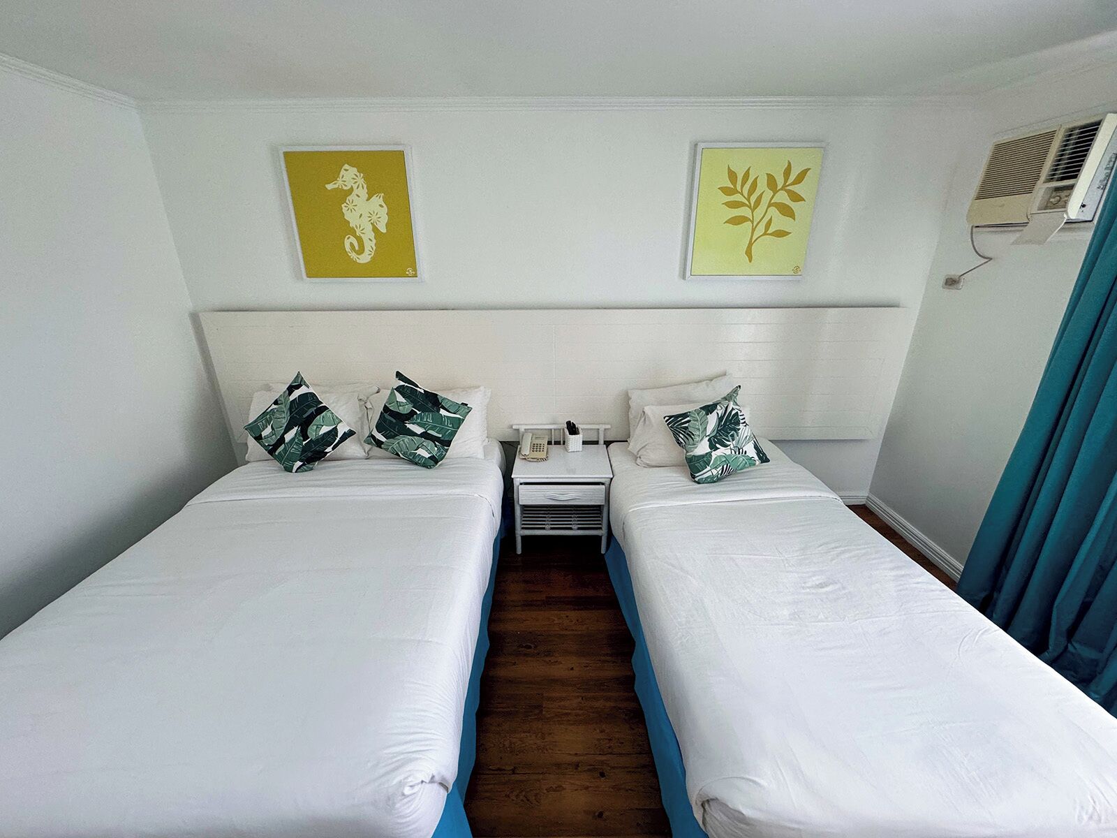 Une chambre d’hôtel avec deux lits simples, du linge de lit blanc et des coussins sur le thème des tropiques. On y trouve une petite table de chevet avec un téléphone et une lampe. Les murs sont de couleur unie et ornés de deux éléments décoratifs.