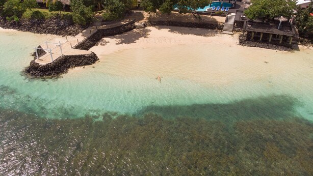 Vista aérea de un complejo turístico en la playa con aguas turquesas cristalinas, una playa de arena y una zona de piscina con sombrillas.
